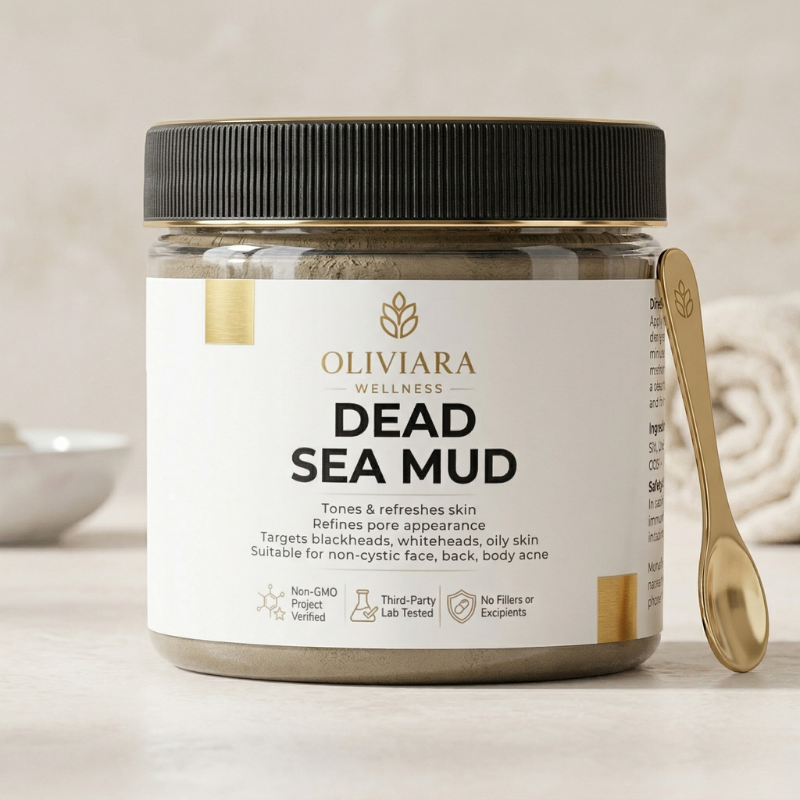 A professional product shot of Oliviara Wellness Dead Sea Mud in a clear jar with a black lid. The jar features a clean white label with gold accents. A gold applicator spoon rests against the jar on a light marble surface, with a blurred bathroom towel in the background.