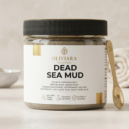 A professional product shot of Oliviara Wellness Dead Sea Mud in a clear jar with a black lid. The jar features a clean white label with gold accents. A gold applicator spoon rests against the jar on a light marble surface, with a blurred bathroom towel in the background.